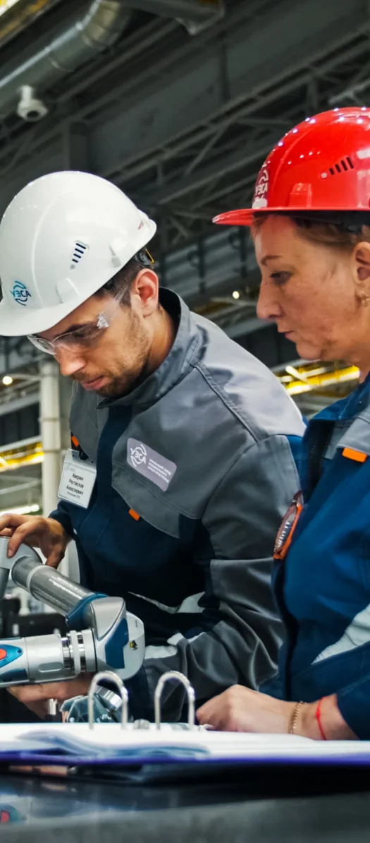 Two factory workers in hard hats and safety glasses looking at a binder beside a machine part