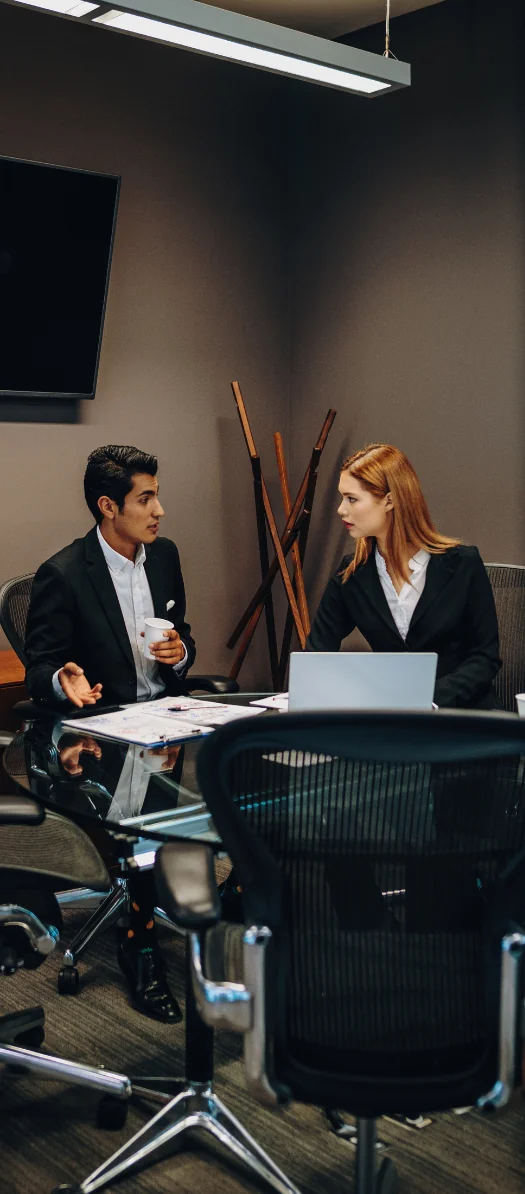 Two professionals talking at a conference table with a laptop and papers